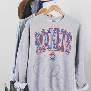 May include: A heather gray sweatshirt with the word "ROCKETS" in blue letters with a red outline. Below the word is a small rocket ship graphic. The sweatshirt is hanging on a wooden hanger.