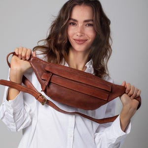 May include: A brown leather fanny pack with a long adjustable strap and a buckle closure. The bag has a rectangular shape with a front pocket and a zipper closure. The fanny pack is held up by a person wearing a white button-down shirt.