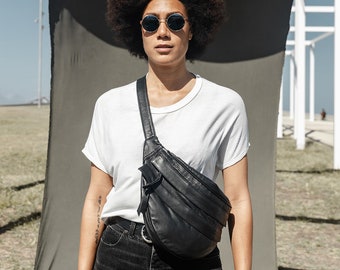 Op de afbeelding: Een persoon draagt een wit T-shirt, een zwarte jeans en een zwart leren heuptasje. Ze draagt ronde zonnebrillen en heeft een grote afro.