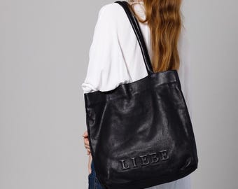 May include: Black leather tote bag with the word "LIEBE" embossed on the front. The bag has two shoulder straps and is carried by a person wearing a white shirt and blue jeans.