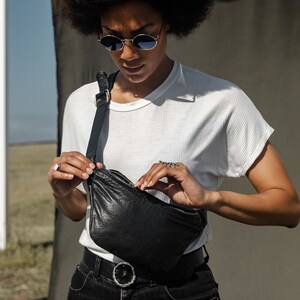 Puede incluir: Un bolso de cintura de cuero negro con una correa larga ajustable. La persona lleva una camiseta blanca, vaqueros negros y gafas de sol. El fondo es de color neutro.