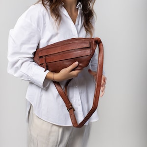 May include: A brown leather fanny pack with a long adjustable strap. The bag has two zippered compartments and a buckle closure. The fanny pack is held in front of a person wearing a white button-down shirt and beige pants.