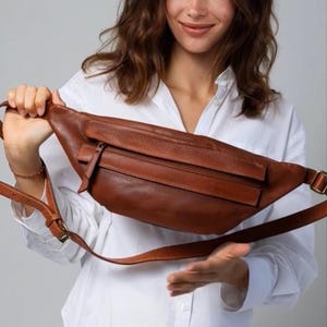 May include: A brown leather fanny pack with a zippered front pocket and adjustable strap. The bag is a warm, medium brown color and has a classic, minimalist design. The strap features a metal buckle.