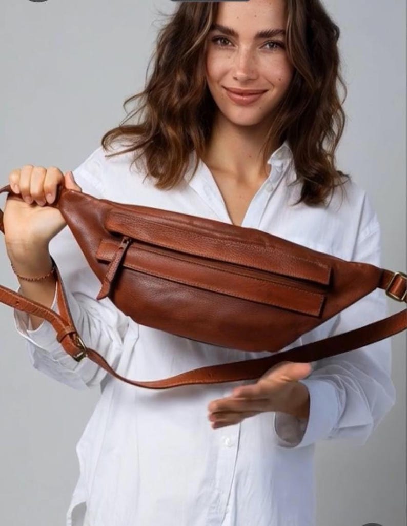 May include: A brown leather fanny pack with a zippered front pocket and adjustable strap. The bag is a warm, medium brown color and has a classic, minimalist design. The strap features a metal buckle.