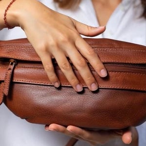 May include: A brown leather fanny pack with a zipper closure and a strap. The bag is held by a person wearing a white shirt. The fanny pack is a rich, warm brown color.