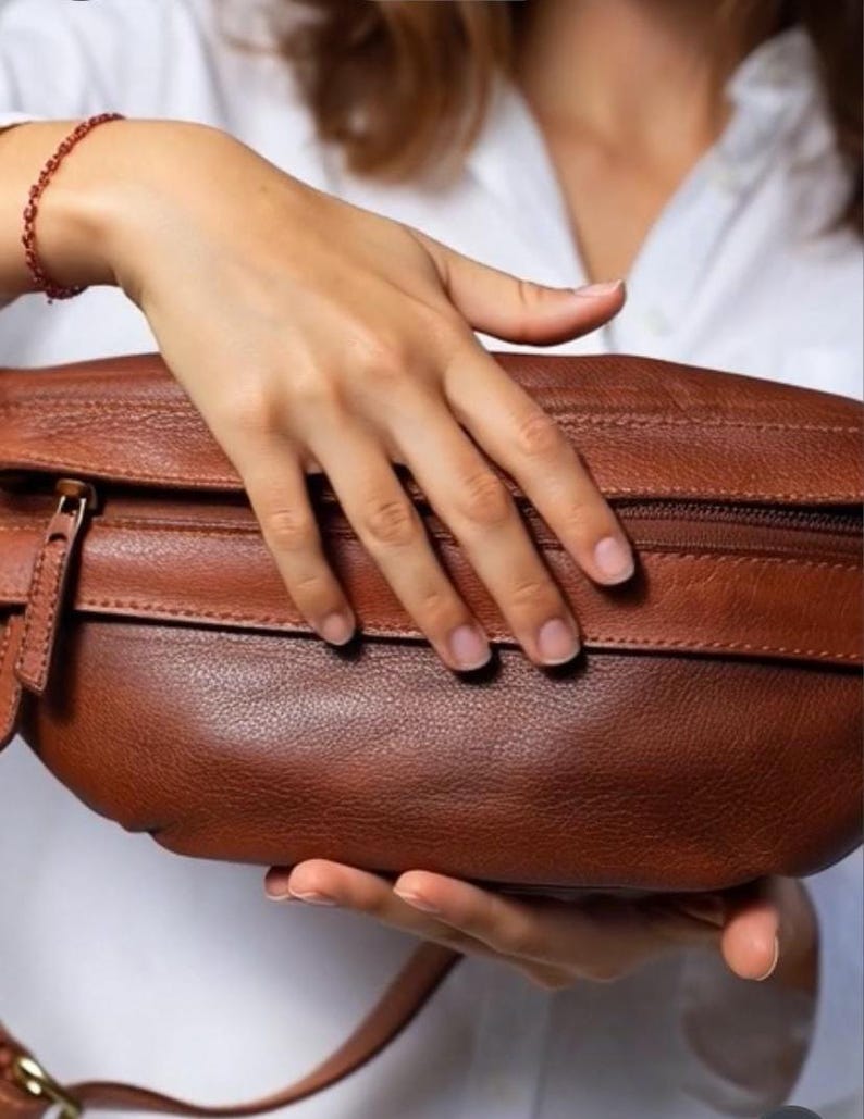 May include: A brown leather fanny pack with a zipper closure and a strap. The bag is held by a person wearing a white shirt. The fanny pack is a rich, warm brown color.