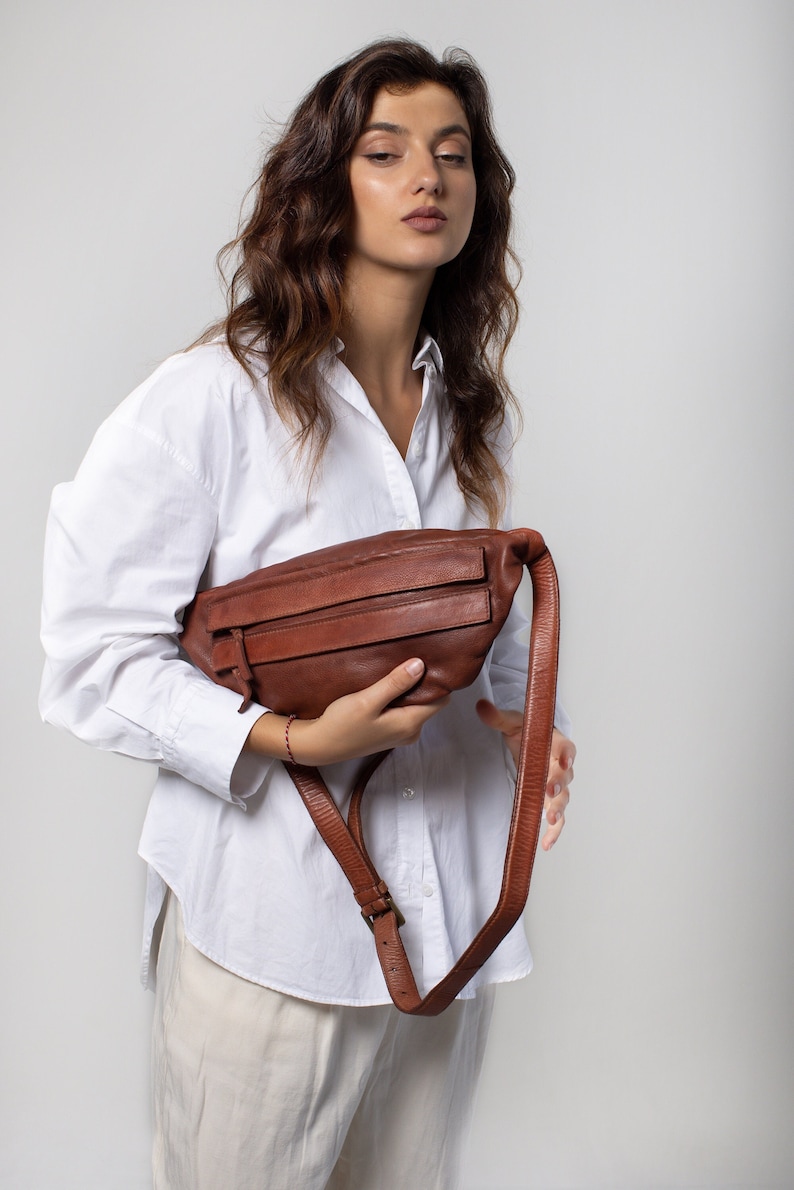 May include: A brown leather fanny pack with a long adjustable strap. The bag has two zippered compartments and a buckle closure. The fanny pack is held in front of a person wearing a white button-down shirt and beige pants.