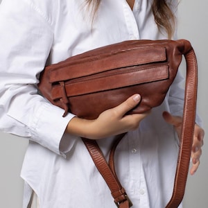 May include: A brown leather fanny pack with two front zippered pockets and a long adjustable strap. The bag is held in front of a person wearing a white button-down shirt.
