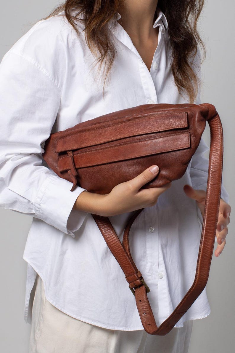 May include: A brown leather fanny pack with two front zippered pockets and a long adjustable strap. The bag is held in front of a person wearing a white button-down shirt.
