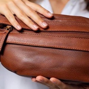 May include: A brown leather bag with a textured surface. The bag has a zipper closure and a small strap. The bag is held by a person.