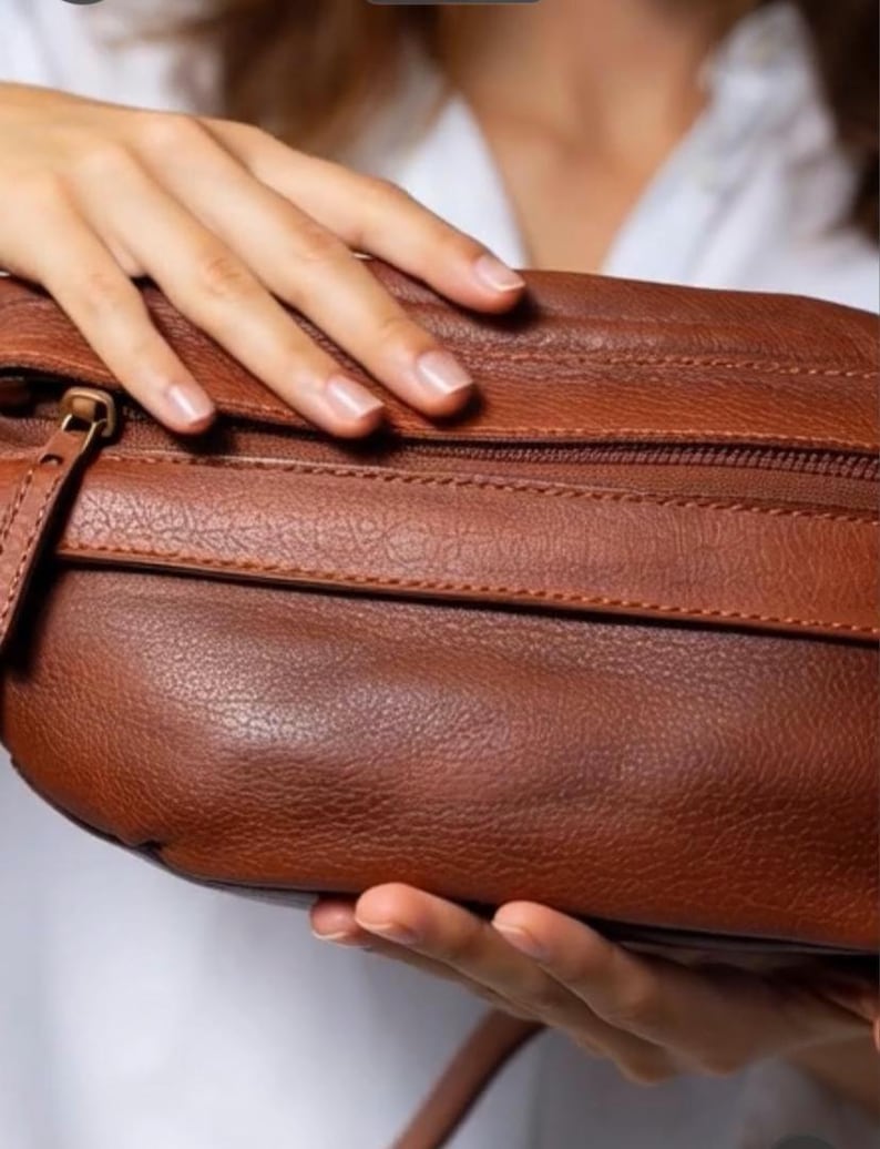 May include: A brown leather bag with a textured surface. The bag has a zipper closure and a small strap. The bag is held by a person.