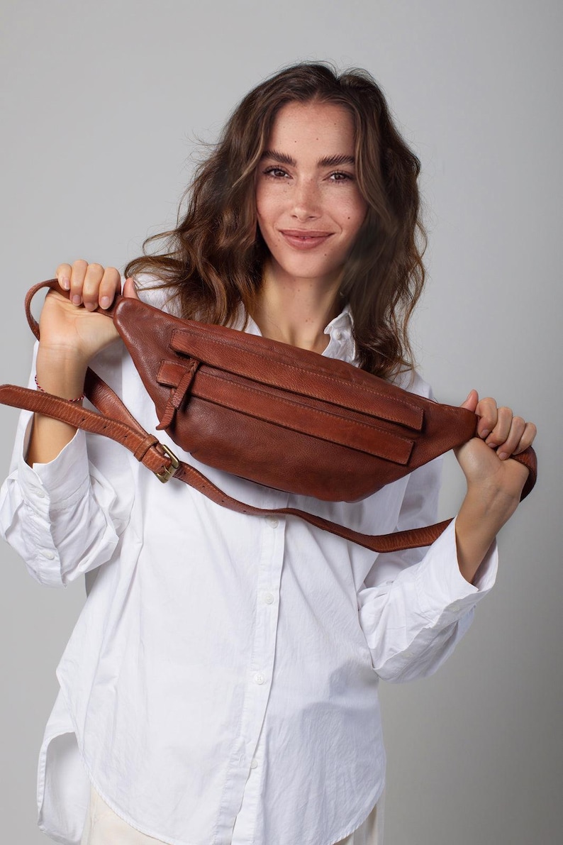 May include: A brown leather fanny pack with a long adjustable strap and a buckle closure. The bag has a rectangular shape with a front pocket and a zipper closure. The fanny pack is held up by a person wearing a white button-down shirt.