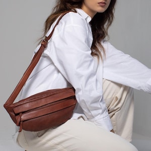 May include: A brown leather crossbody bag with a long strap. The bag has a rectangular shape with a flap closure and horizontal stitched details. The bag is worn over the shoulder.