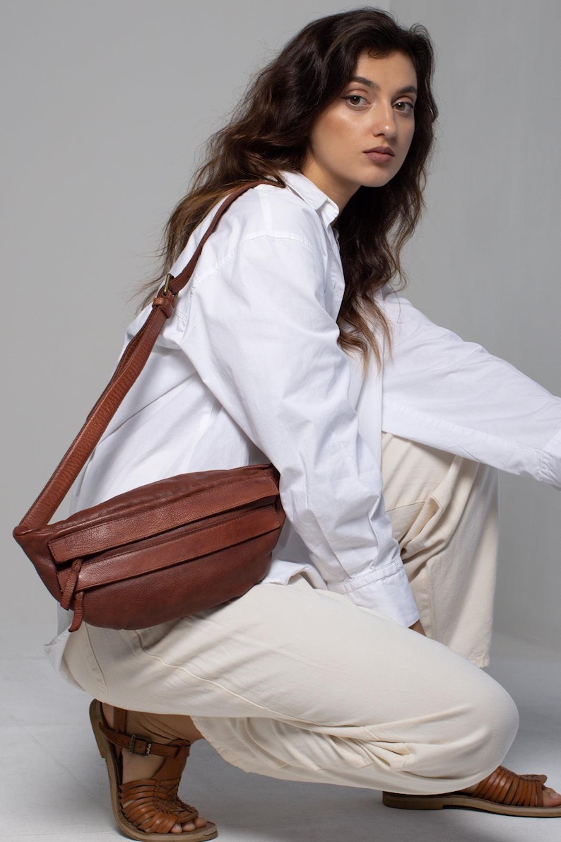 May include: A brown leather crossbody bag with a long strap. The bag has a rectangular shape with a flap closure and horizontal stitched details. The bag is worn over the shoulder.