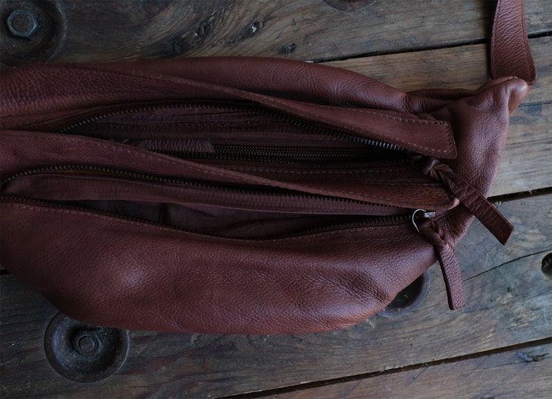 May include: A brown leather fanny pack with multiple zippered compartments. The bag is open, revealing the interior lining. The leather has a textured appearance, and the zippers are dark brown. The bag is designed to be worn around the waist.