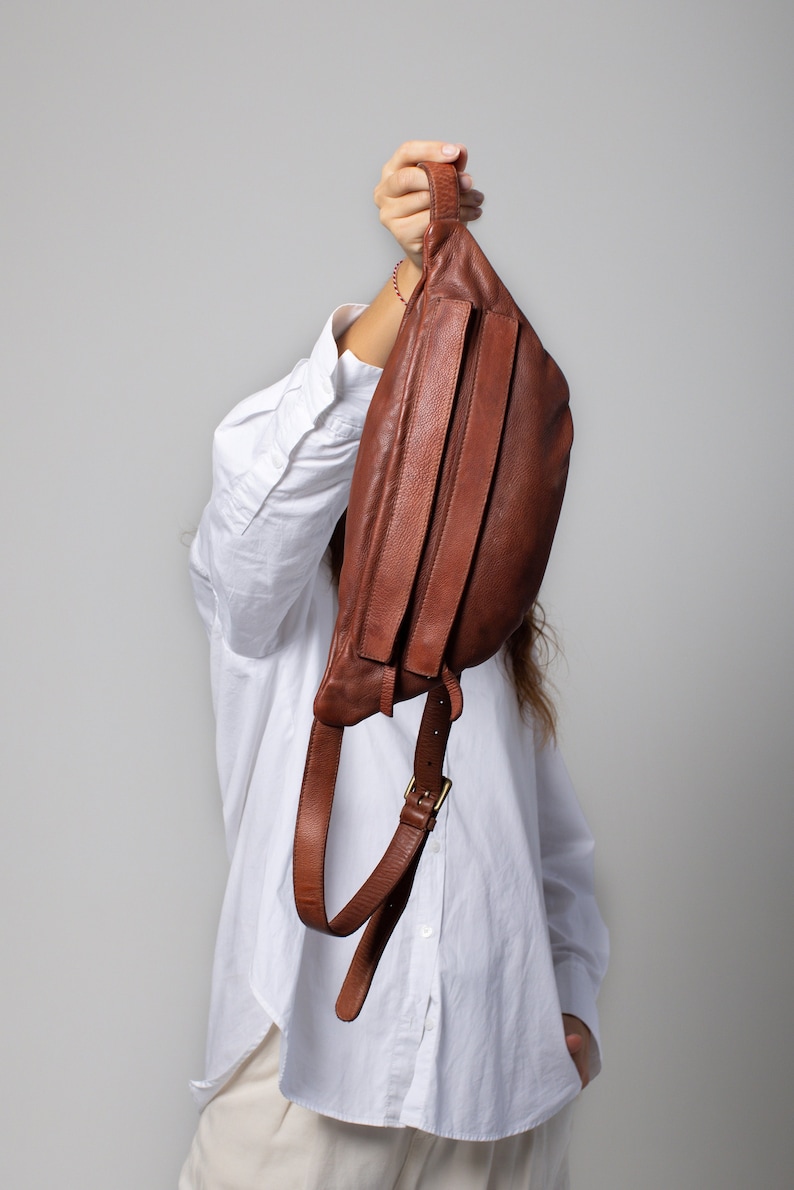May include: A brown leather fanny pack with a curved shape and adjustable strap. The bag has a buckle closure and a handle at the top. The leather appears to be textured, and the bag is held up by a person.