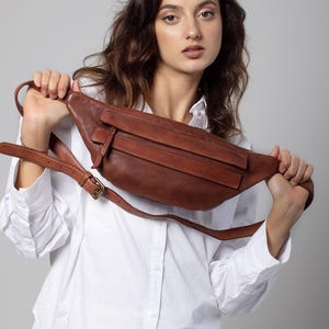 May include: A brown leather fanny pack with a long adjustable strap and a buckle closure. The bag has a rectangular shape with a front flap and a zippered pocket. The fanny pack is held up by a person wearing a white button-down shirt.