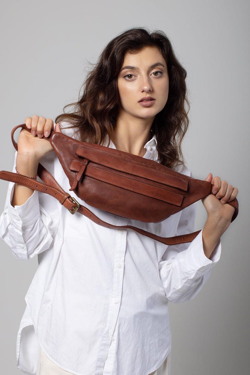 May include: A brown leather fanny pack with a long adjustable strap and a buckle closure. The bag has a rectangular shape with a front flap and a zippered pocket. The fanny pack is held up by a person wearing a white button-down shirt.
