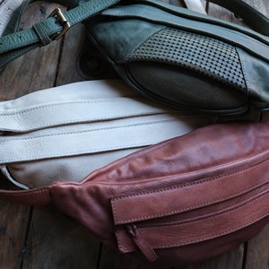 May include: Three leather fanny packs in shades of green, cream, and brown. The green pack has a perforated design. Each pack has a strap and a zipper closure. The bags are displayed on a wooden surface.
