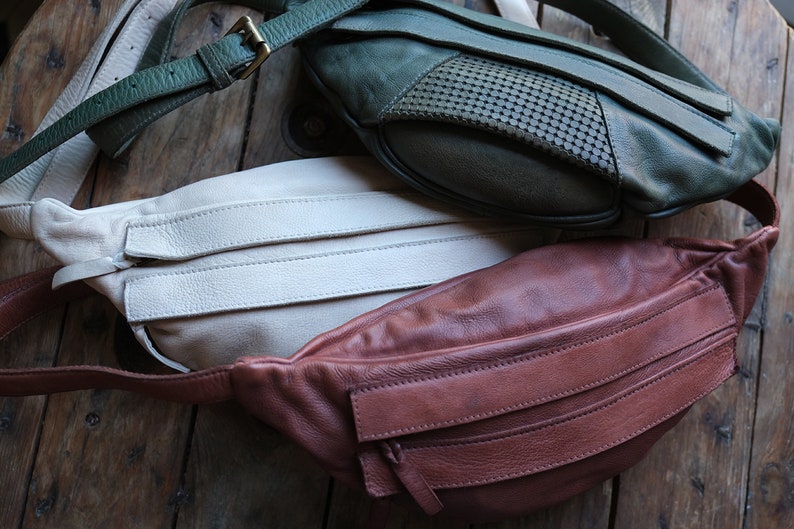 May include: Three leather fanny packs in shades of green, cream, and brown. The green pack has a perforated design. Each pack has a strap and a zipper closure. The bags are displayed on a wooden surface.