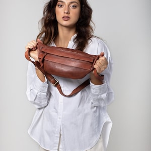 May include: A brown leather fanny pack with a gold-colored buckle. The bag has a long adjustable strap and a zippered main compartment. The fanny pack is held up by a person wearing a white button-down shirt and cream-colored pants.