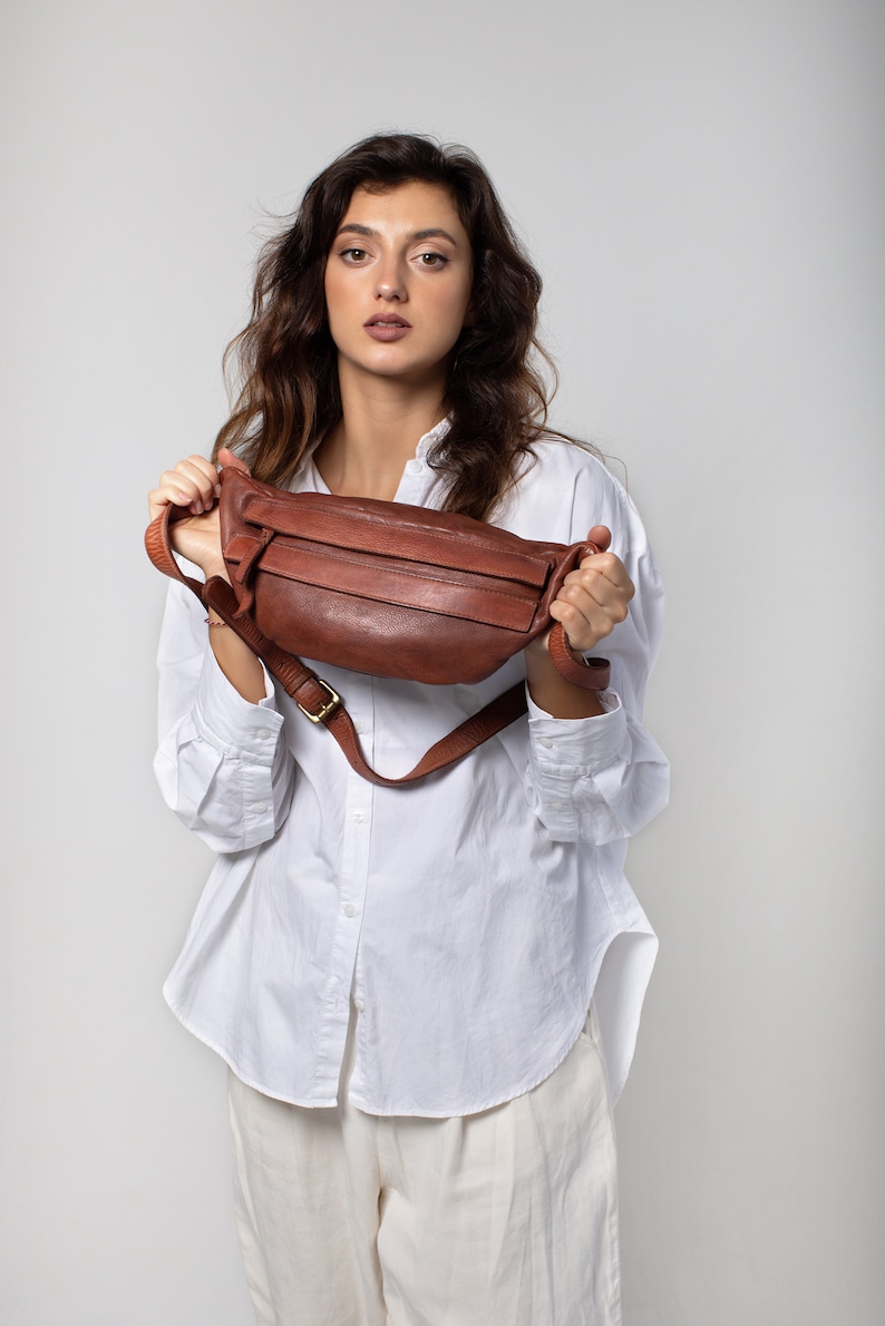 May include: A brown leather fanny pack with a gold-colored buckle. The bag has a long adjustable strap and a zippered main compartment. The fanny pack is held up by a person wearing a white button-down shirt and cream-colored pants.