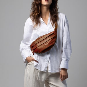 May include: A brown leather fanny pack with a zipper closure and a strap. The bag is worn across the body. The person is wearing a white button-down shirt and cream-colored pants.
