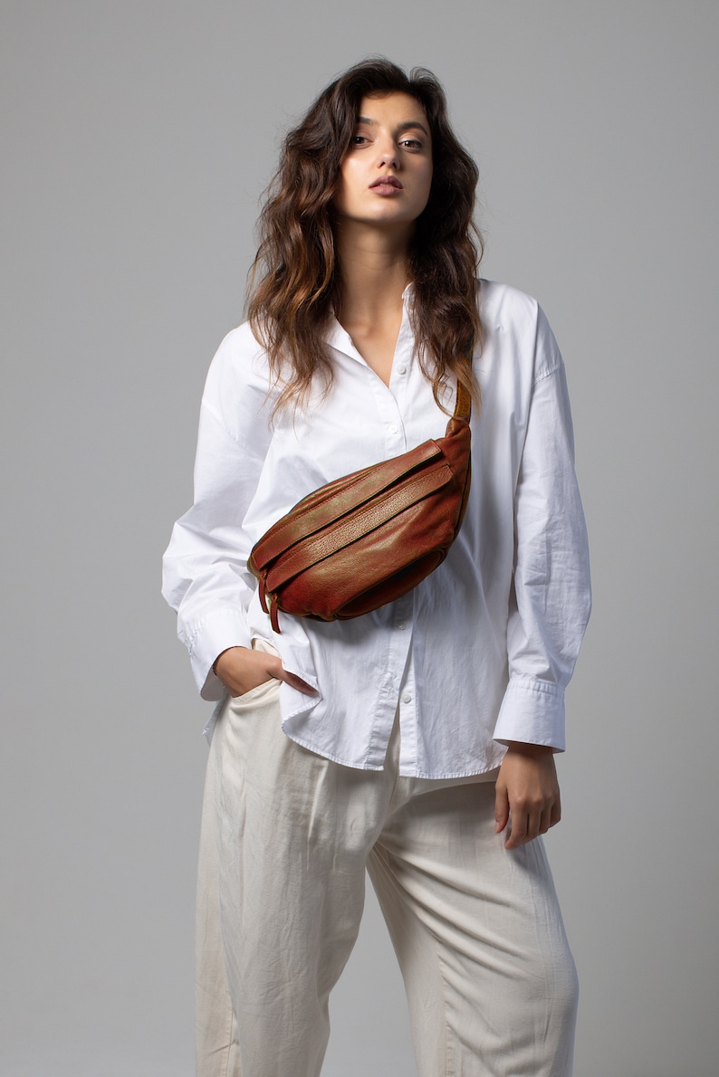 May include: A brown leather fanny pack with a zipper closure and a strap. The bag is worn across the body. The person is wearing a white button-down shirt and cream-colored pants.