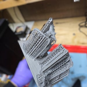 May include: A gray 3D-printed object, possibly a model or prototype, held by a gloved hand. The object has a complex, geometric structure with multiple layers and intricate details. The print is on a rectangular base.
