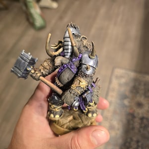May include: A detailed figurine of a fantasy creature, possibly a minotaur, holding a large hammer. The figure has fur, horns, and armor with purple accents. The hammer is gray, and the base is a rocky terrain.