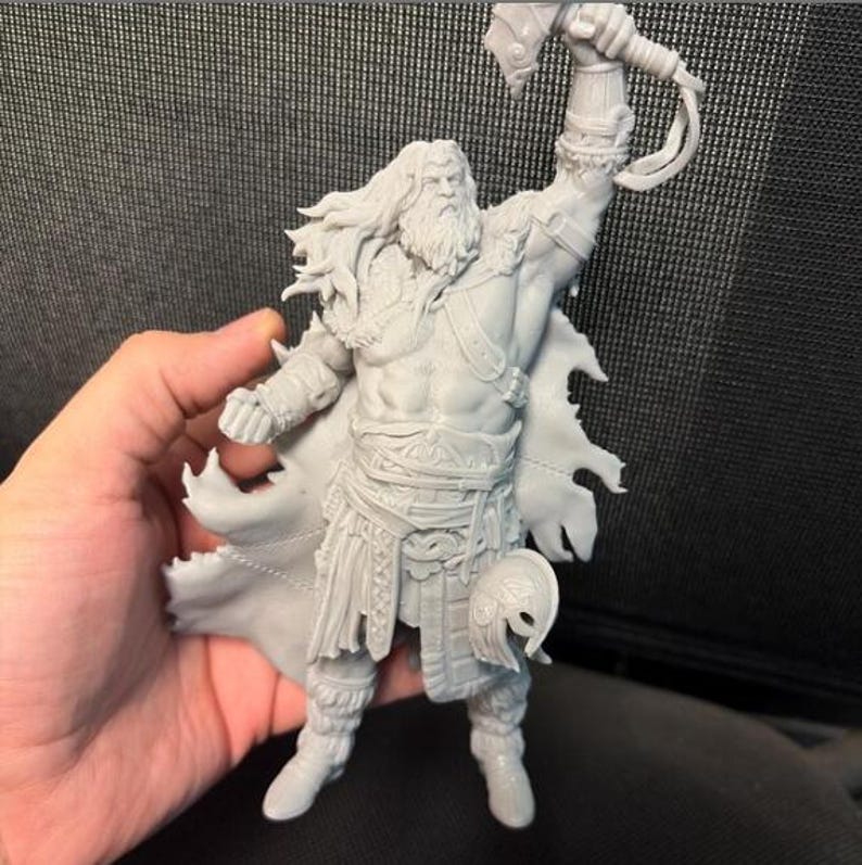以下が含まれることがあります： 筋肉質でひげを生やした男性の灰色のフィギュアで、戦士のポーズでハンマーを高く掲げています。このフィギュアは詳細な鎧、マントを身に着けており、長い髪をしています。ヘルメットが足元にあります。フィギュアは手で持たれています。