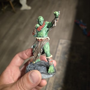 May include: A hand-painted miniature figurine of a green-skinned orc in a fighting stance. The orc is wearing red and brown armor and is standing on a gray stone base. The figurine is held in a person's hand.