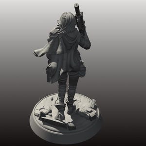 以下が含まれることがあります： 未来的な衣装を着た女性キャラクターの灰色のフィギュアで、ライフルを持っています。フィギュアは、散らばった破片のある円形のベースに立っています。キャラクターはフード付きのマントと戦術的なギアを着用しています。