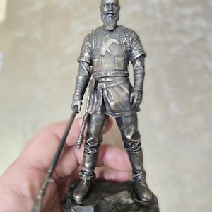 以下が含まれることがあります： 槍を持った、ひげを生やした戦士の銀色の置物。このフィギュアは詳細な鎧を身につけ、岩の台座の上に立っています。戦士の服装にはチュニック、ズボン、ブーツが含まれます。槍は左手に持たれています。