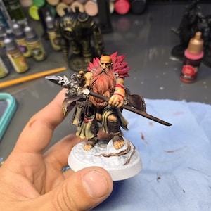 May include: A miniature fantasy figure of a bearded warrior with a red beard and feathered shoulder pads. The figure is holding a spear and is mounted on a white base. The figure is painted with detailed colors.