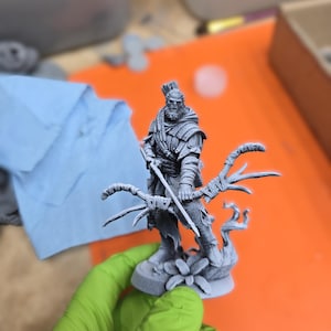 以下が含まれることがあります： 弓と矢を持った戦士の灰色のミニチュアフィギュア。フィギュアは花の模様の台座の上に立っています。弓は枝でできています。戦士は鎧とマントを着ています。