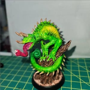 以下が含まれることがあります： 手描きのミニチュアフィギュア。角、トゲ、長い赤い舌を持つ緑色のトカゲのような生き物。フィギュアは茶色のテクスチャベースに配置され、詳細で鮮やかな塗装が施されています。