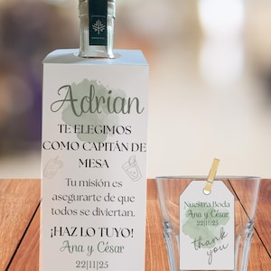 Puede incluir: Una botella de vidrio transparente con tapa dorada y una caja blanca con el nombre "Adrian" y otro texto. Un vaso transparente con una etiqueta que dice "Nuestra Boda Ana y César 22/11/25 thank you" también está en la imagen.
