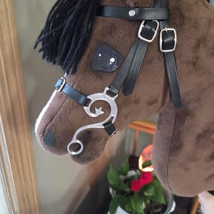 May include: A brown plush horse head with a black mane and a black leather bridle. The bridle features silver buckles and a decorative bit. The toy horse head is soft and plush.