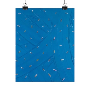 May include: A poster print featuring an aerial view of numerous colorful surfboards scattered across a vibrant blue ocean. The surfboards vary in color, including yellow, pink, and white, creating a dynamic and visually appealing scene. The poster is held by two black binder clips.