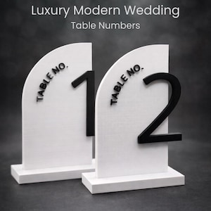 Pode incluir: Sinais brancos de números de mesa com os números pretos 1 e 2. Os sinais têm uma base retangular e um topo curvo. O texto "TABLE NO." é impresso no canto superior esquerdo do sinal. O texto "Luxury Modern Wedding Table Numbers" está no topo.