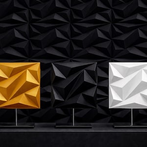 以下が含まれることがあります： 金、黒、白の3つの幾何学的な正方形の彫刻。それぞれ細い金属製のスタンドに黒いベースで取り付けられています。背景は、繰り返しの三角形のパターンを持つテクスチャ加工された黒い壁です。