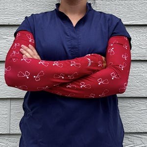 May include: A person wearing a navy blue scrub top with red arm sleeves. The sleeves are patterned with white stethoscopes. The person's arms are crossed in front of them.