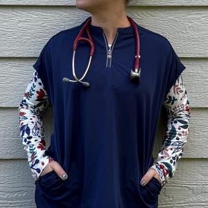 May include: Navy blue medical scrub top with a quarter-zip neckline and floral patterned sleeves. The sleeves feature a white background with red, green, and blue floral designs. A red stethoscope hangs around the neck.
