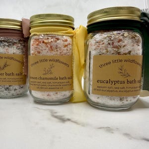 May include: Three glass jars of bath salts with gold lids and labels. The labels read "lavender bath salt", "lemon chamomile bath salt", and "eucalyptus bath salt". Each jar is filled with a mixture of salts and dried botanicals.