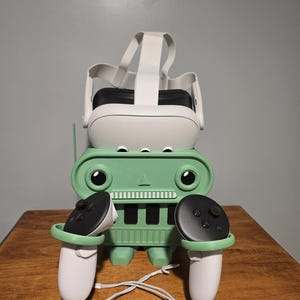 May include: A green robot-shaped VR headset stand holding a white VR headset and two white controllers with black buttons. The stand has a cartoonish design with circular eyes and a rectangular body. The controllers are connected by a white cable.