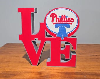 May include: A red and gray "LOVE" sign with a Phillies logo. The sign features the word "LOVE" in large, block letters, with the "O" replaced by a circular logo with the team name and a Liberty Bell.