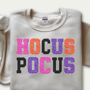May include: A beige sweatshirt with the words "HOCUS POCUS" in a colorful, glittery font. The top row of text is pink, orange, black, and purple. The bottom row is black, purple, pink, and purple.