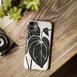 May include: A white phone case featuring a black leaf design. The case has a matte finish and is shown on a wooden surface. The design is a stylized illustration of a large leaf.