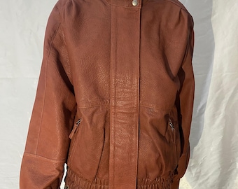 May include: A reddish-brown leather jacket with a high collar and a zipper closure. The jacket has a ribbed hem and two front pockets with zippers. The sleeves are long and have a slightly puffed design at the shoulders.